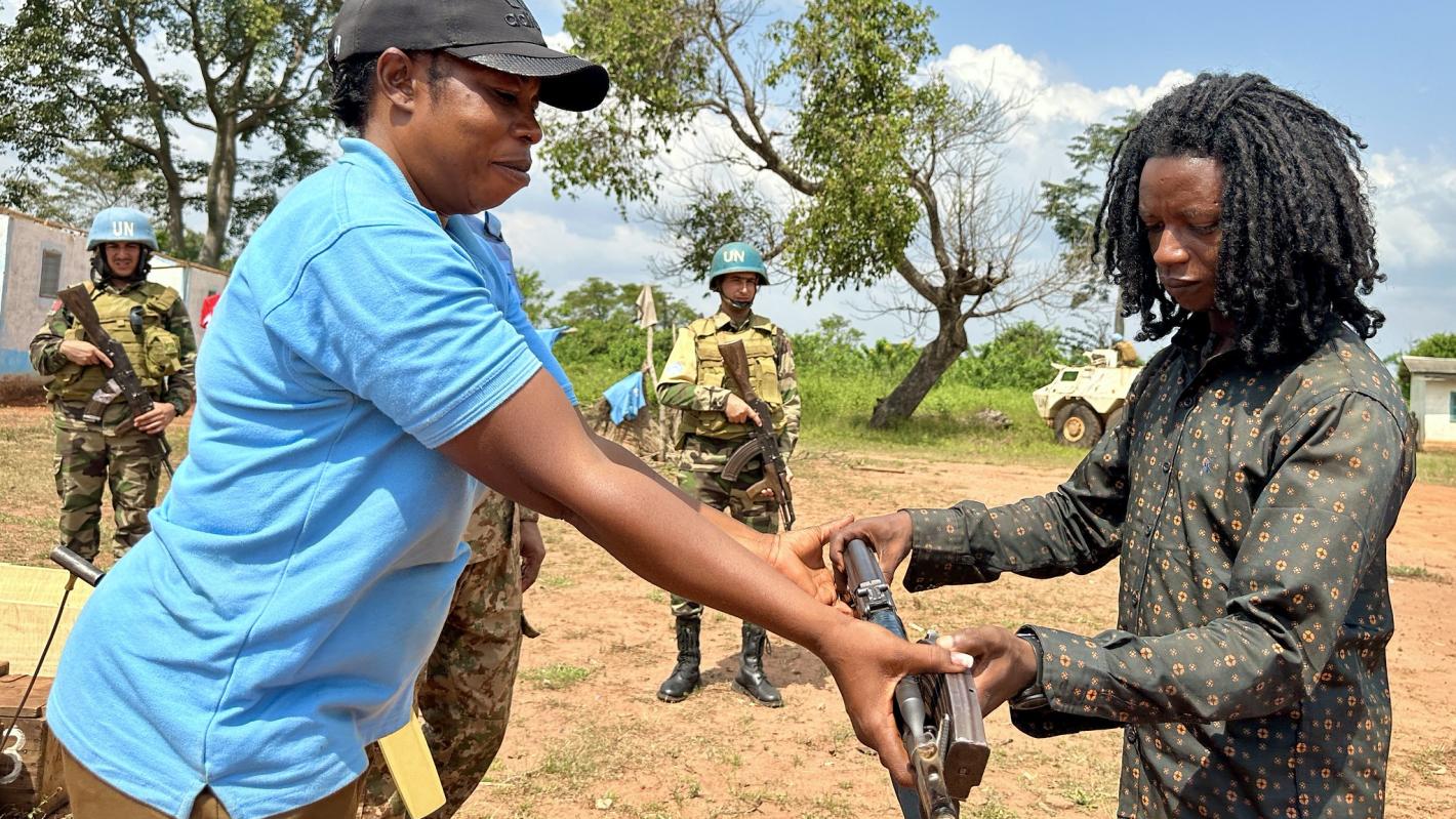 Two individuals outdoors exchanging a rifle, with one person wearing a blue shirt and cap and the other in a patterned shirt. In the background, uniformed personnel and a white vehicle are visible near trees under a partly cloudy sky.