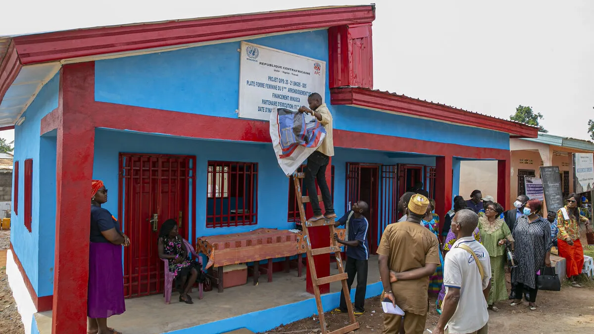 La mairie du 6è arrondissement de Bangui a réceptionné, lors d’une cérémonie, le mardi 7 décembre 2021, un bâtiment construit dans le cadre des projets à impact rapide, pour la plateforme féminine. Ce bâtiment entièrement équipé qui comprend quatre pièces, dont deux bureaux, a été financé par la MINUSCA A man on a ladder unveils a sign on a new single-story building in front of a crowd