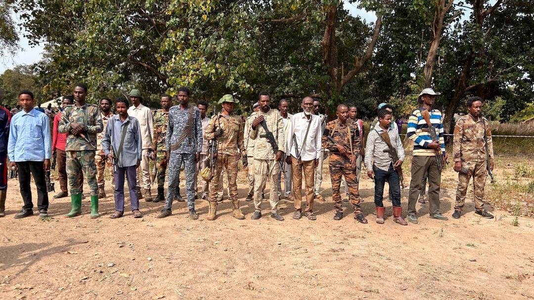 A group of people standing in a line outdoors on dry ground, with trees in the background. Several individuals are wearing military-style camouflage uniforms, while others are dressed in casual clothing. Some are holding rifles.