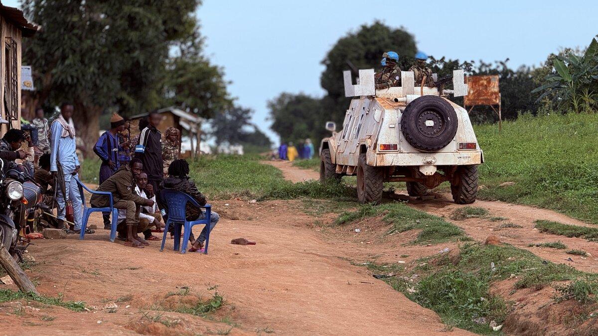 Un véhicule de maintien de la paix de l’ONU passe devant un groupe de personnes assises dehors.