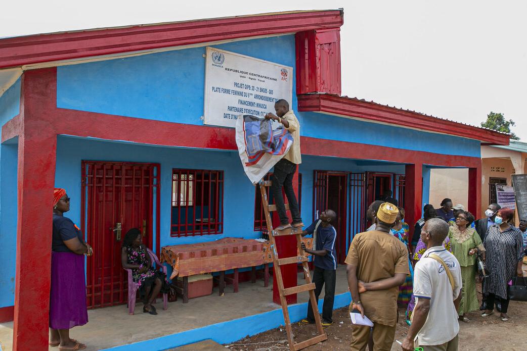 A man on a ladder unveils a sign on a new single-story building in front of a crowd