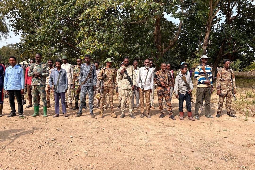 A group of people standing in a line outdoors on dry ground, with trees in the background. Several individuals are wearing military-style camouflage uniforms, while others are dressed in casual clothing. Some are holding rifles.