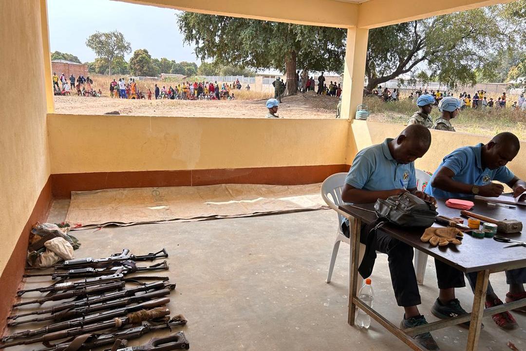 Two individuals seated at a table inside a covered area, working with documents and items. Several rifles are laid out on the floor nearby. Outside, a group of people and uniformed personnel can be seen in an open area with trees.