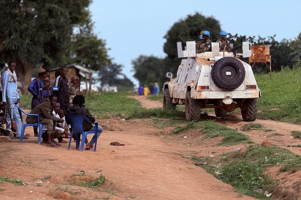 Un véhicule de maintien de la paix de l’ONU passe devant un groupe de personnes assises dehors.