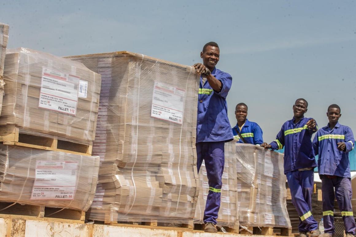 Four workers wearing blue uniforms with reflective stripes stand on a flatbed truck loaded with large pallets wrapped in plastic. Each pallet has visible labels, and the setting appears to be outdoors under a clear sky.