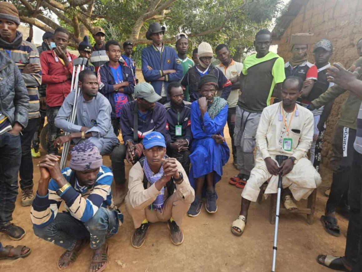Un groupe d'hommes se tiennent debout et accroupis à l'extérieur, sous un arbre.