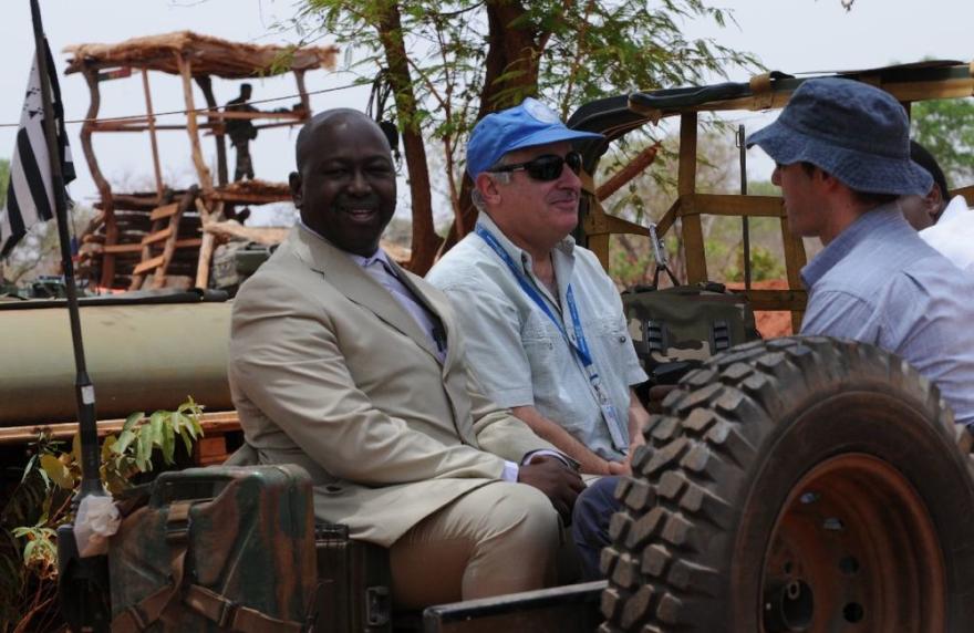 Two individuals sit in an open military vehicle with UN markings; a wooden lookout structure and trees are in the background.